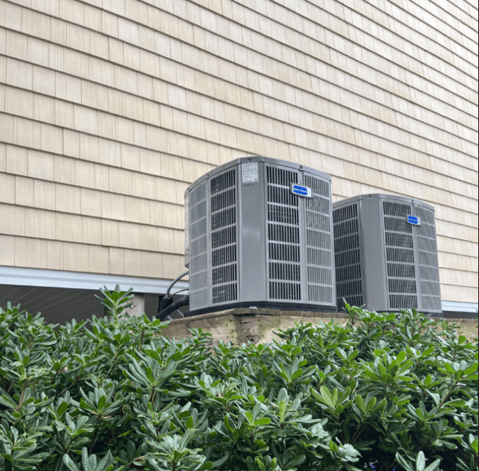Two outdoor air conditioning units are mounted on a wooden platform next to a house with tan siding, above green shrubs.